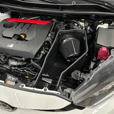 Close-up of the engine bay of a Toyota GR Yaris. The focus is on the installed RamAir Proram intake kit. Visible are the air filter, the heat shield, and the intake manifold. The kit is neatly installed and fits well into the engine bay.