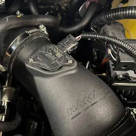 Close-up of the RamAir Proram intake kit for the Toyota GR Yaris. The black plastic intake pipe with sensor is installed. The RamAir logo is clearly visible. Various cables and hoses in the background.