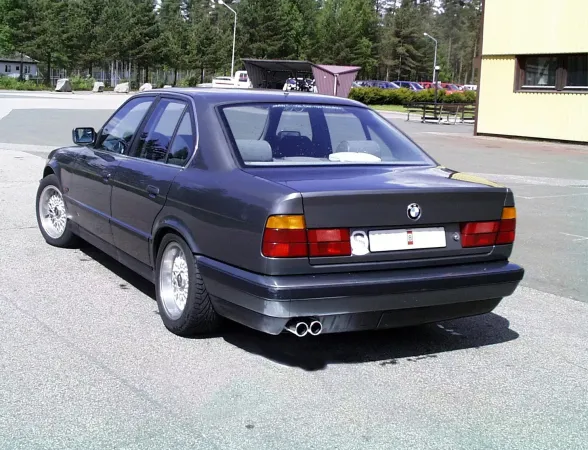 Rear view of a dark gray BMW E34. You can see the chrome steel duplex sports exhaust system with 2x70mm tailpipes, installed below the bumper. The vehicle is standing in a parking lot, with trees and a building in the background.